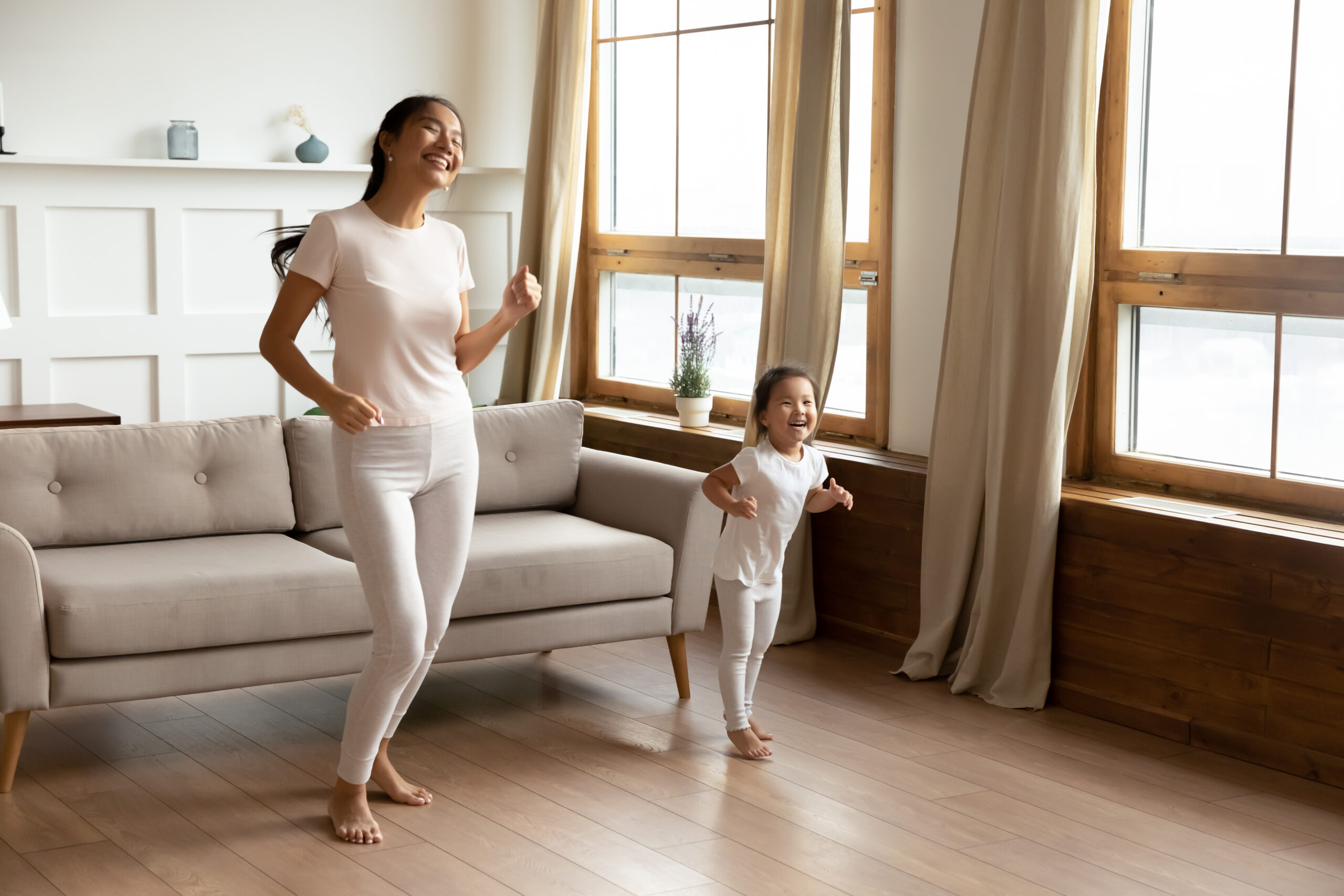 Full length asian happy mother little daughter in casual comfort clothes standing barefoot on warm wooden floor in sunny living room enjoy life, listen music dancing, having fun at modern home concept